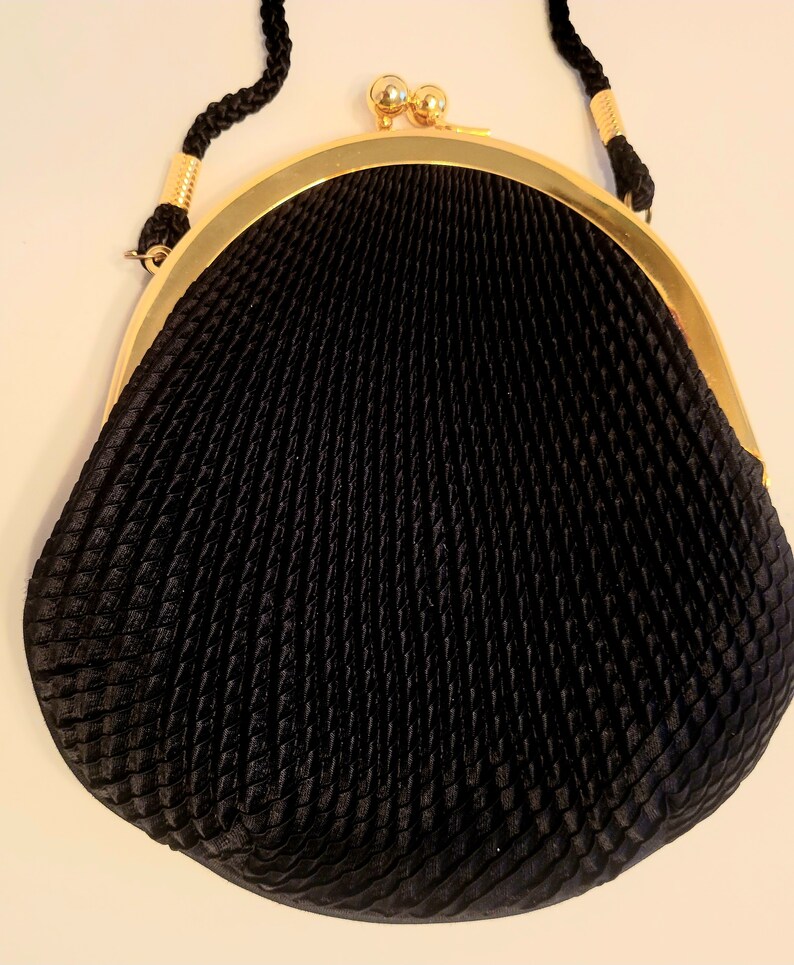 May include: Black evening bag with a textured, pleated design. The bag features a gold-tone frame and clasp, with a braided black strap. The bag's shape is rounded at the bottom and tapers towards the top, creating a classic silhouette.