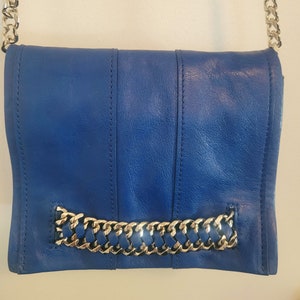 Puede incluir: Un bolso bandolera de cuero azul cobalto con una correa de cadena plateada. El bolso presenta un detalle de cadena plateada en la parte delantera y costuras verticales. El bolso es de color liso y tiene un cierre de solapa.