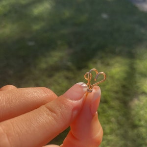 May include: A gold-colored heart-shaped charm with a delicate design. The charm is held by a finger.