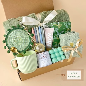 May include: A gift box containing a sage green textured blanket with a white ribbon, a green pom-pom coaster, a gold tea infuser, a light green ribbed mug with a gold spoon, pens, a shea butter tube, a green floral notebook, and a bubble candle.