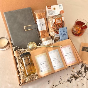 May include: A gift box containing a gray notebook, a package of Frontier Sausage and Lentil soup mix, cinnamon toffee, a tin of Restored candle, a gold tea infuser, a jar of honey, a wooden honey dipper, and three tea pouches labeled Morning Summit, Quiet Whisper, and English Breakfast Tea.
