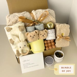 May include: A gift box containing a beige textured blanket with a gold ribbon, a cream muslin cloth with bear prints, a crochet rabbit rattle, bear-faced socks, a yellow ribbed mug, a tea infuser, a candle, tea leaves, and a card with text.