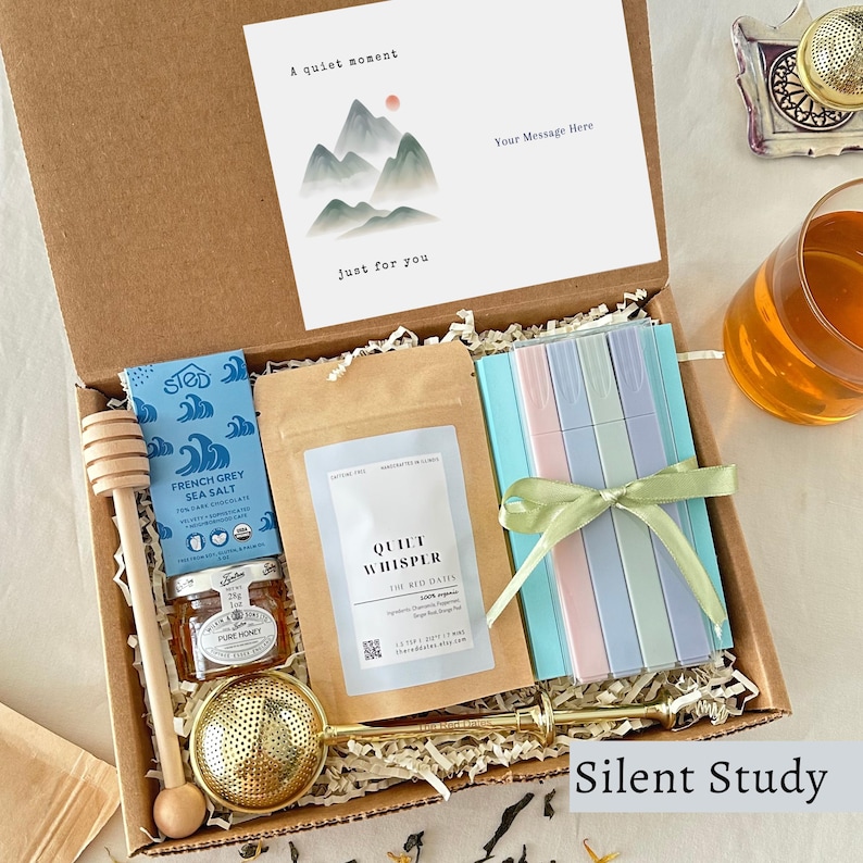 May include: A gift box containing a wooden honey dipper, a jar of honey, a bar of French Grey Sea Salt dark chocolate, a pouch of Quiet Whisper tea, a set of pastel markers tied with a green ribbon, and a card with mountain art and text.