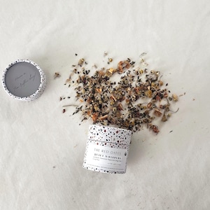 May include: A cylindrical tea container with a white and multicolored terrazzo pattern. The label reads THE RED DATES QUIET WHISPERS. Loose leaf tea is spilled beside the container. The lid is separate, showing the text Have a Tea-riffic day.