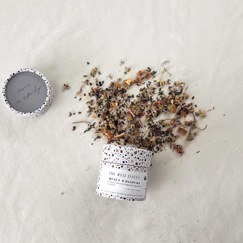 May include: A cylindrical tea container with a white and multicolored terrazzo pattern. The label reads THE RED DATES QUIET WHISPERS. Loose leaf tea is spilled beside the container. The lid is separate, showing the text Have a Tea-riffic day.