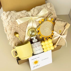 May include: A gift box containing a beige fuzzy blanket with a cream ribbon, a yellow ribbed mug with a gold spoon, a jar of The Red Dates lemon meringue honey, a gold tea infuser, a yellow pom-pom coaster, a bubble candle, and a wooden honey dipper.
