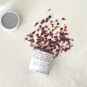 May include: A round, speckled container of loose-leaf tea, labeled "The Red Dates Hibiscus Bloom," with scattered tea leaves and berries. The lid of a matching container is nearby, with the text "Have a tea-riffic day!"