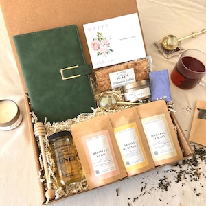 May include: A gift box filled with a green journal, tea, honey, a candle, and a card that says "Happy Mother's Day". The box includes tea bags labeled "Hibiscus Glow", "Lemon Meringue", and "Morning Summit".