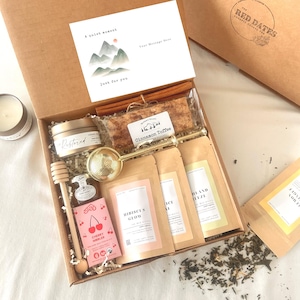 May include: A gift box filled with tea, honey, toffee, and a candle. The box includes a card with the text "A quiet moment, just for you". The box also contains tea bags labeled "Hibiscus Glow" and "Bland Breeze".