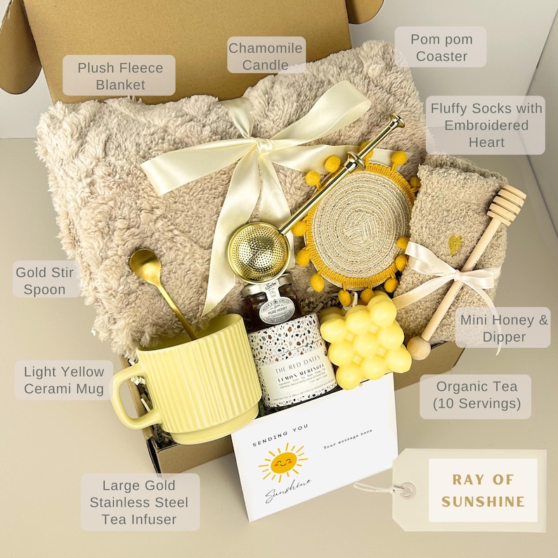 May include: A gift box containing a beige plush fleece blanket with a cream ribbon, a light yellow ceramic mug, a gold stir spoon, a gold stainless steel tea infuser, a chamomile candle, a pom pom coaster, fluffy socks, and honey with a dipper.