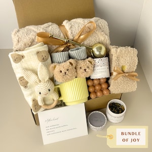 May include: A gift box containing a beige textured blanket with a gold ribbon, a cream muslin cloth with bear prints, a crochet rabbit rattle, bear-faced socks, a yellow ribbed mug, a tea infuser, a candle, tea leaves, and a card with text.