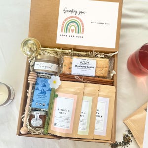 May include: A gift box containing a gold tea infuser, a wooden honey dipper, a jar of honey, a tin of tea, a package of blueberry lemon biscotti, cinnamon sticks, and three pouches of tea labeled Hibiscus Glow, Spring Mint, and Earl Grey Lavender.