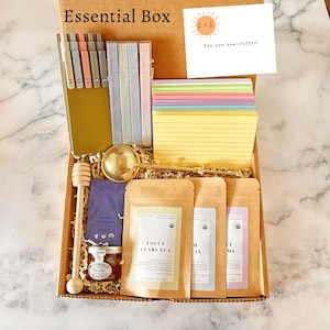 May include: A cardboard box labelled "Essential Box" containing stationery, tea, and honey. Includes pens, notepads, tea bags, a honey dipper, and a card that says "You are tea-riffic!"