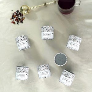 May include: Six cylindrical tea canisters with a white and black terrazzo pattern, each featuring a label reading The Red Dates. A gold-toned metal tea infuser, a small pile of loose tea leaves, and a glass mug filled with dark tea are also present.