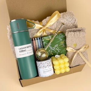 May include: A gift box containing a 500ml green vacuum bottle, a set of pens, a gold tea infuser, a green floral pouch, a yellow bubble candle, a small container labeled The Red Dates, and a beige fuzzy blanket tied with a gold ribbon.