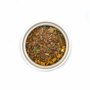 May include: Overhead view of a white ceramic bowl filled with a mix of dried herbs and spices. The blend includes green leaves, brown cinnamon sticks, and orange pieces. The bowl is centered on a white background.