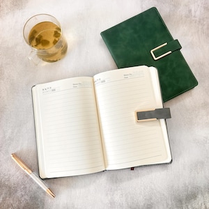May include: An open, lined notebook with a gold pen and a closed green notebook with a gold clasp. A glass of tea sits nearby. The open notebook has the words "Memo No. Date" printed at the top of each page.