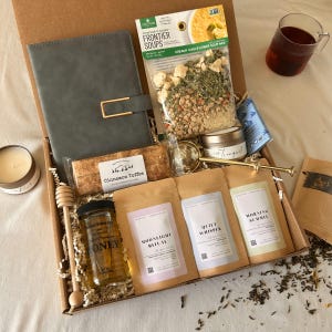 May include: A gift box containing a gray notebook, a bag of Frontier Soups Creamy Cauliflower Soup Mix, a jar of honey with a wooden dipper, Cinnamon Toffee biscuits, three tea pouches labeled Moonlight Ritual, Quiet Whisper, and Morning Summit, a gold tea infuser, and a small candle.