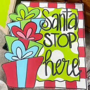 May include: A Christmas-themed wooden sign with the words "Santa STOP here" in black script. The sign features a green polka-dot background and a border of red and white stripes. Decorative gift boxes are stacked on the left side.