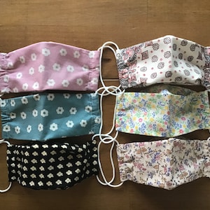 May include: Six different fabric face masks with various floral patterns. The masks are all white with different colored flowers and designs. The masks are all handmade and have elastic ear loops.