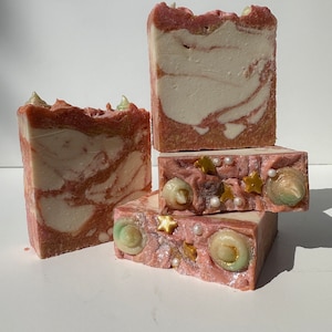 May include: A stack of four handmade soap bars with a pink and white marbled design. The top bar has a swirl design with gold glitter and green and white embellishments.