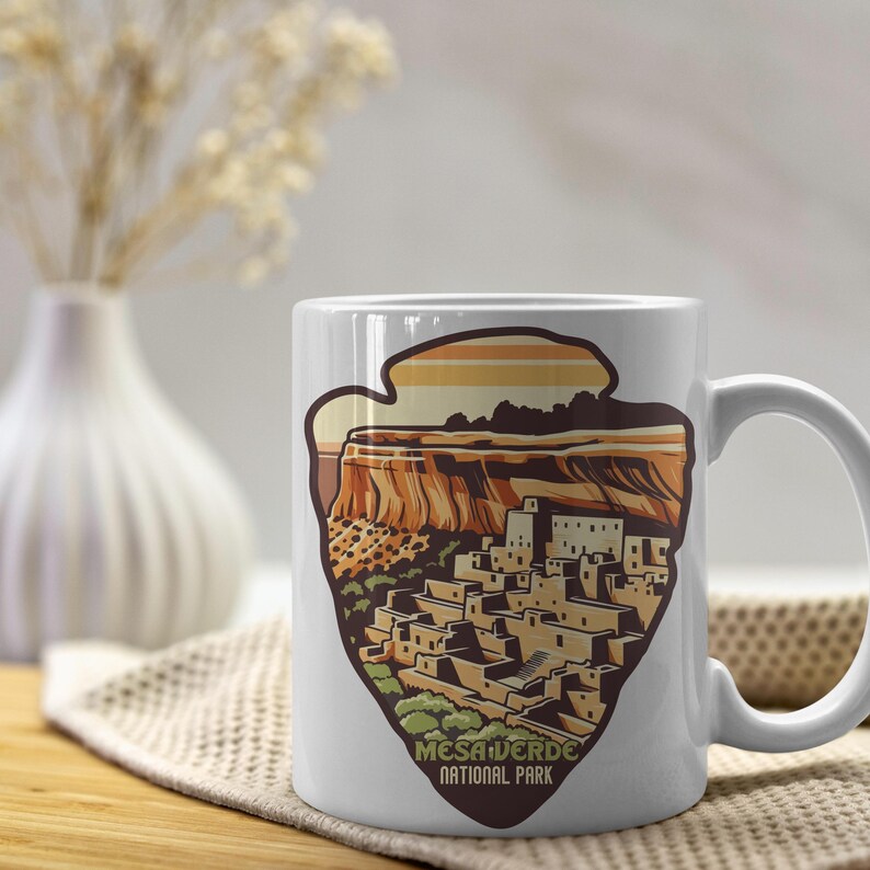 Puede incluir: Taza de cer&aacute;mica blanca con una ilustraci&oacute;n colorida del Parque Nacional Mesa Verde. La ilustraci&oacute;n presenta una vivienda en un acantilado con una puesta de sol en el fondo. El texto "Mesa Verde National Park" est&aacute; impreso debajo de la ilustraci&oacute;n.