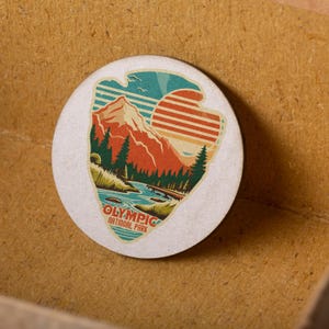 Puede incluir: Una pegatina redonda blanca con una ilustraci&oacute;n de estilo vintage de una cordillera, un r&iacute;o y una puesta de sol. El texto "OLYMPIC NATIONAL PARK" est&aacute; impreso debajo de la ilustraci&oacute;n.
