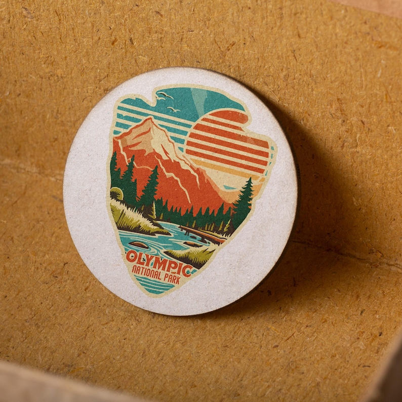 Puede incluir: Una pegatina redonda blanca con una ilustraci&oacute;n de estilo vintage de una cordillera, un r&iacute;o y una puesta de sol. El texto "OLYMPIC NATIONAL PARK" est&aacute; impreso debajo de la ilustraci&oacute;n.