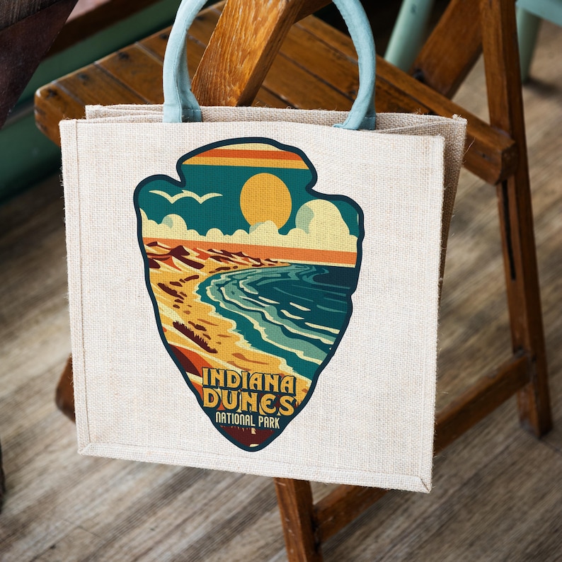 Puede incluir: Una bolsa de tela beige con un asa azul. La bolsa presenta una ilustraci&oacute;n de estilo vintage de una escena de playa con dunas de arena, olas del oc&eacute;ano y un sol. El texto "Indiana Dunes National Park" est&aacute; impreso debajo de la ilustraci&oacute;n.