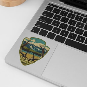 Puede incluir: Una pegatina verde, marr&oacute;n y azul con un dise&ntilde;o vintage que presenta una cordillera, un lago y una pareja de cuernos. La pegatina dice "Gates of the Arctic National Park".