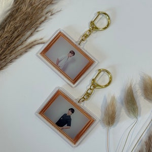 May include: Two clear acrylic keychains with gold chains. Each keychain features a photo of a person in a framed picture. The frames are brown with a gold border.