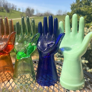 May include: Four colorful glass hand-shaped decorative objects. The hands are in shades of orange, green, blue, and light green. They are displayed outdoors on a metal table with a natural background.