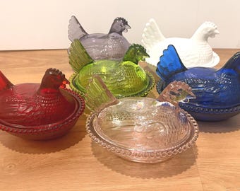 May include: A collection of six glass hen-shaped lidded dishes in various colors including red, lime green, cobalt blue, clear, white, and a translucent grey. Each dish features a textured feather pattern and a beaded rim on the base.