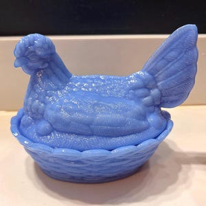 May include: A light blue hen-shaped trinket box. The hen is perched atop a basket-like base. The box is made of a solid, opaque material with a textured surface. The hen's feathers and the basket's woven design are detailed.