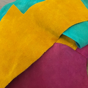 May include: Close-up of colorful suede leather pieces in shades of teal, gold, and magenta. The textures vary, with some pieces cut into irregular shapes. The leather appears soft and pliable, suitable for various crafting projects.