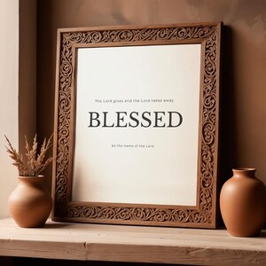 May include: A wooden framed print with the word "BLESSED" in bold black letters. The frame has an ornate carved design. The text "The Lord gives and the Lord takes away, be the name of the Lord" is above and below the word "BLESSED".