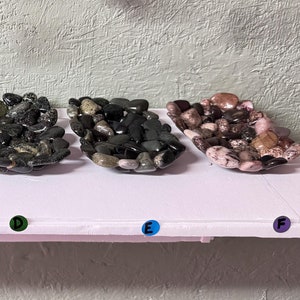 May include: Five small trays filled with various colored smooth stones. Each tray is labeled with a letter from A to E. Above the trays, five stones are hanging from chains, also labeled A to E.