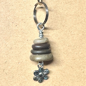 May include: A keychain featuring a silver-toned metal lobster clasp and a circular split ring. Attached to the ring is a stack of four smooth, rounded, earth-toned stones secured with wire, finished with a small silver-toned metal flower charm.