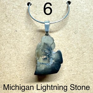 May include: A pendant featuring a Michigan Lightning Stone (Septarian Nodule) in the shape of the state. The stone is approximately 3/4 inch in size and is attached to a silver-colored bail and a silver-colored necklace loop. The number "6" is above the pendant.