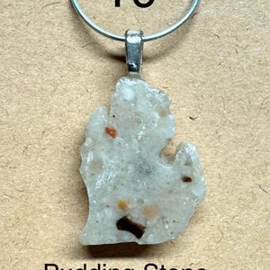 May include: A pendant featuring a Pudding Stone in the shape of the state of Michigan. The stone is light gray with brown and orange inclusions, and is approximately 1 inch in length. The pendant hangs from a silver-colored metal loop.