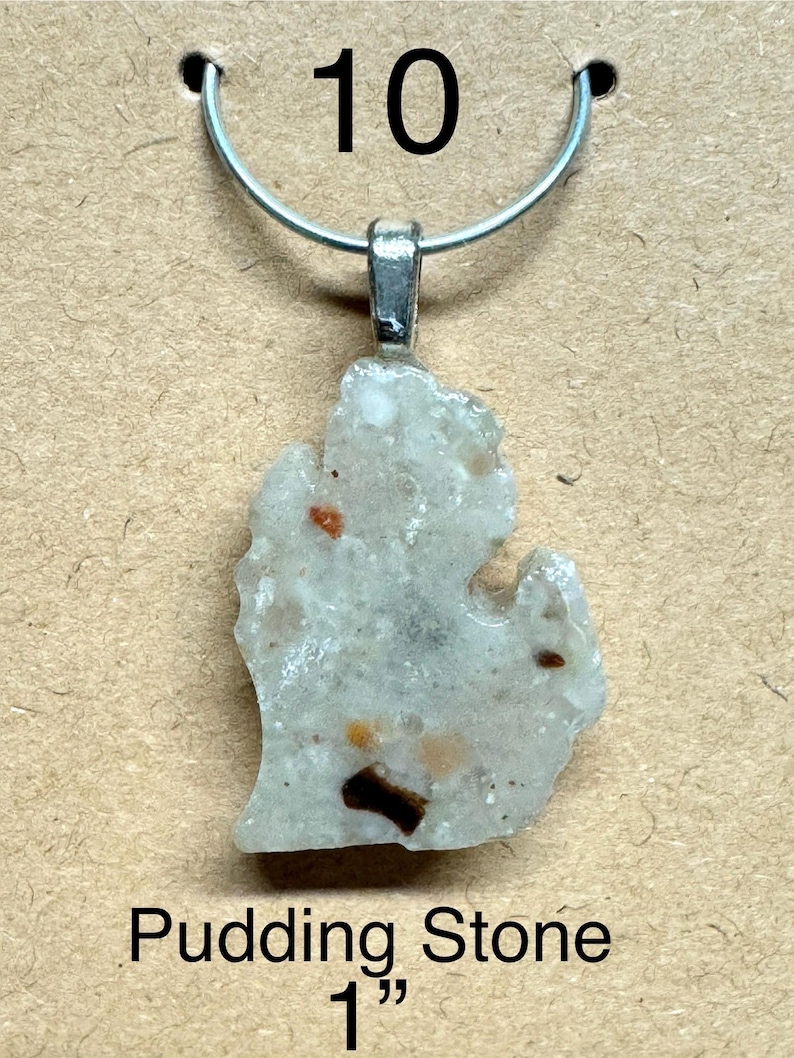 May include: A pendant featuring a Pudding Stone in the shape of the state of Michigan. The stone is light gray with brown and orange inclusions, and is approximately 1 inch in length. The pendant hangs from a silver-colored metal loop.