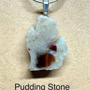 May include: A pendant featuring a Pudding Stone, approximately 1 inch in length. The stone is a light gray color with brown and red inclusions. The pendant hangs from a silver-colored metal loop. The number "12" is above the pendant.