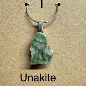 May include: A green and white Unakite pendant in the shape of the state of Michigan, suspended from a silver-colored metal loop. The pendant measures 3/4 inch. The word "Unakite" is printed below the pendant.
