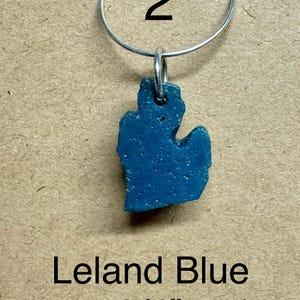 May include: A blue pendant in the shape of the state of Michigan, suspended from a silver-colored metal ring. The pendant is labeled "Leland Blue" and measures 3/4 inch.