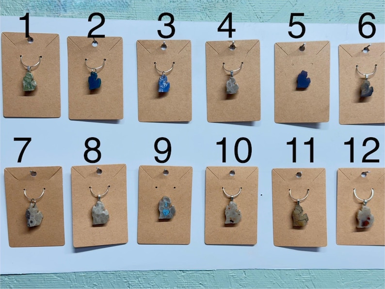 May include: A collection of twelve necklaces, each featuring a pendant shaped like the state of Michigan. The pendants are made of various stones in shades of blue, green, and gray, displayed on individual kraft paper cards.