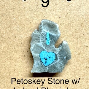 May include: A gray Petoskey stone with a blue heart-shaped inlay and a blue inlay line. The stone is shaped like the state of Michigan. The text "Petoskey Stone w/ Leland Blue inlay 1"" is below the stone.