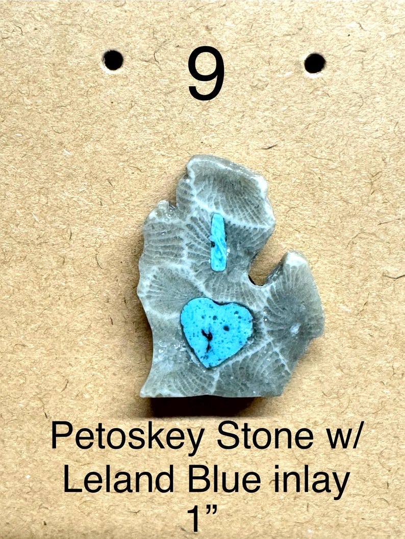 May include: A gray Petoskey stone with a blue heart-shaped inlay and a blue inlay line. The stone is shaped like the state of Michigan. The text "Petoskey Stone w/ Leland Blue inlay 1"" is below the stone.