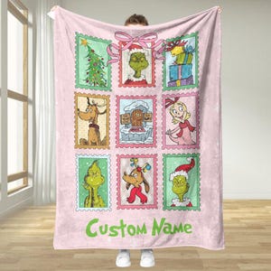 Könnte beinhalten: Eine hellrosa Fleece-Decke mit neun gerahmten Bildern von Charakteren aus einer Weihnachtsgeschichte. Die Bilder zeigen einen Weihnachtsbaum, Geschenke und den Grinch. Die Worte "Custom Name" sind grün gedruckt.