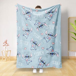 May include: A light blue blanket featuring a repeating pattern of the character Stitch from Lilo & Stitch, holding a pink heart. The word "Stitch" is also printed on the blanket. The blanket is held up by a person.