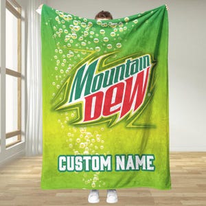 Può includere: Coperta verde con il logo Mountain Dew in verde, rosso e bianco. La coperta ha uno sfondo sfumato con bolle e la scritta "CUSTOM NAME" in bianco.